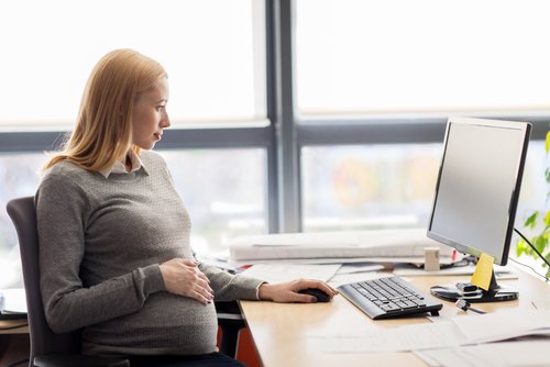 Empleado embarazada sosteniendo su vientre y mirando su computadora en su escritorio en su oficina con una ventana de piso a techo en el fondo