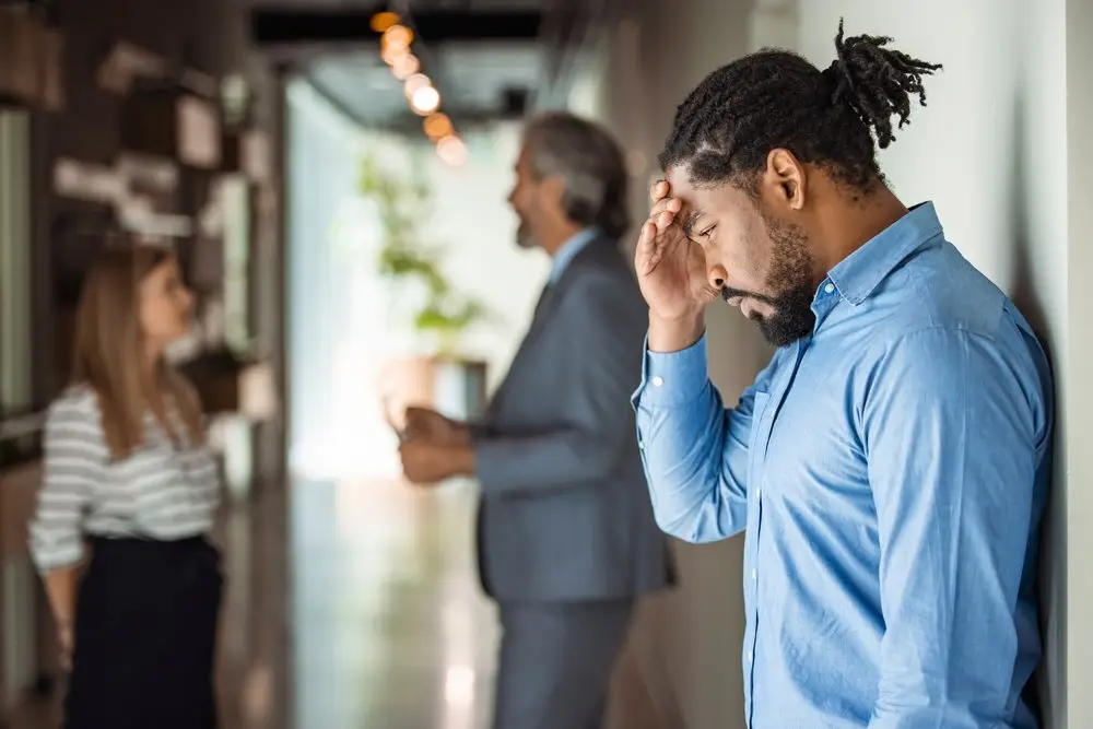 An upset-looking employee leaning against the wall while two co-workers talk in the background