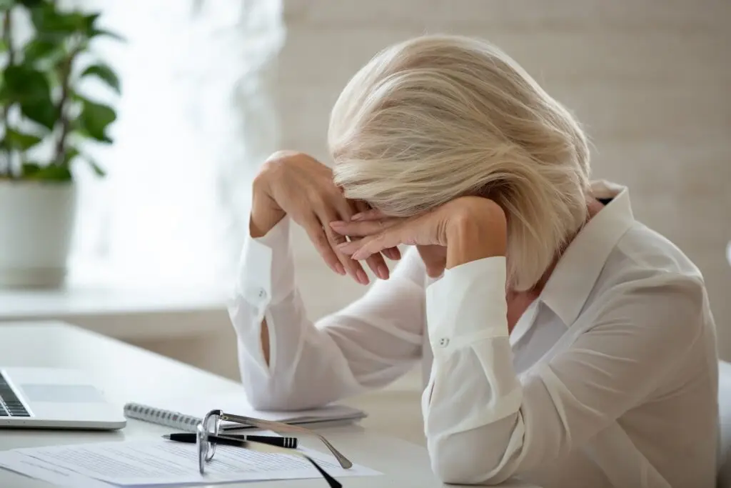 Older woman at her office desk looking distraught and holding her head in her hands with a plant and window in the background