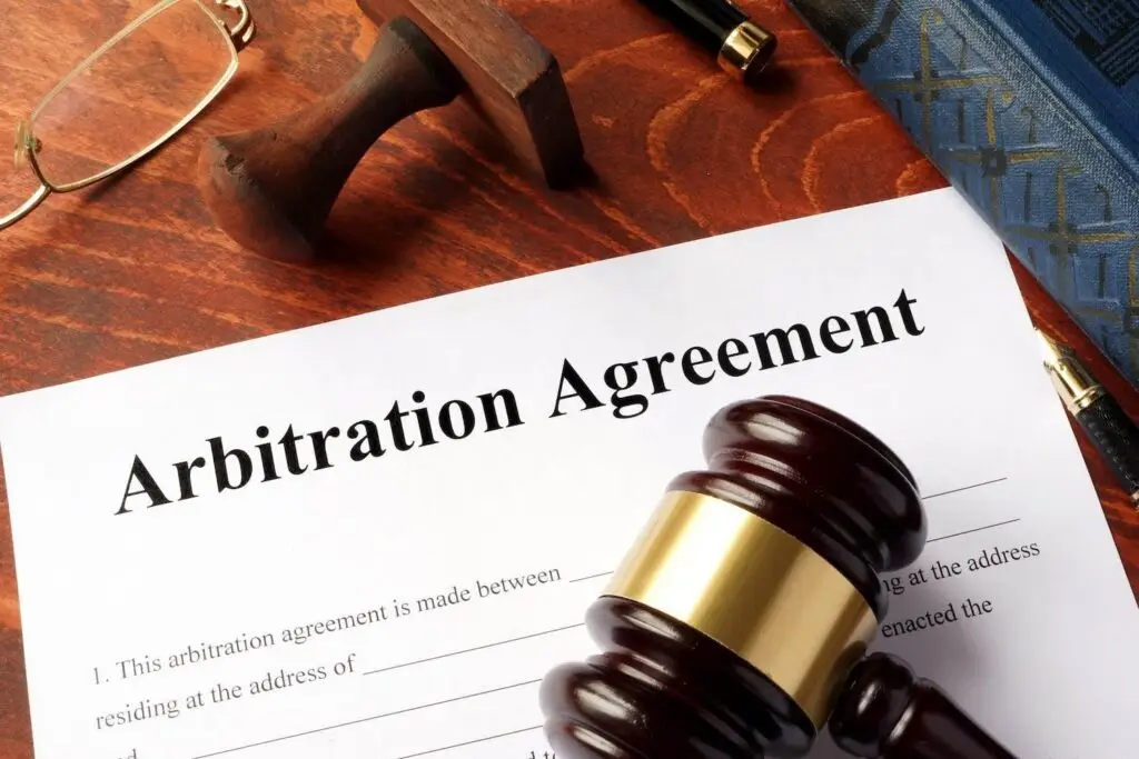 Wooden table upon which there are the following items: A paper with the title "Arbitration Agreement," a wooden gavel, a stamp, a pen, and glasses