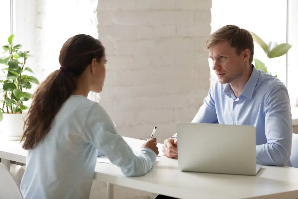 A boss and an employee having a meeting across a white desk. The boss is behind a laptop, and there are plants in the background