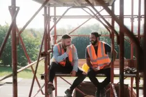 two construction workers in orange vests eating while sitting on scaffolding 