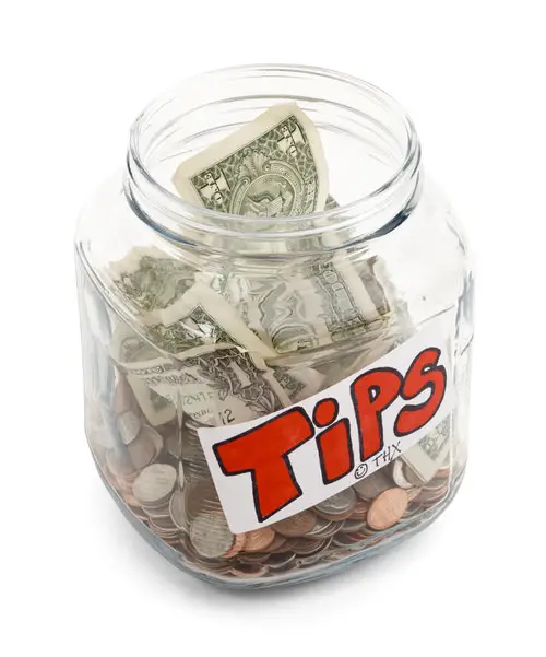 Large glass jar labeled "Tips" in big red letters against a white background, and it contains several dollar bills and coins. 
