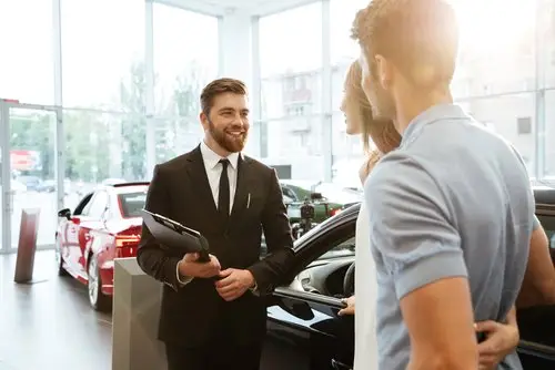 Car salesman in a business suit holding a clipboard greeting a young couple in the showroom of a car dealership