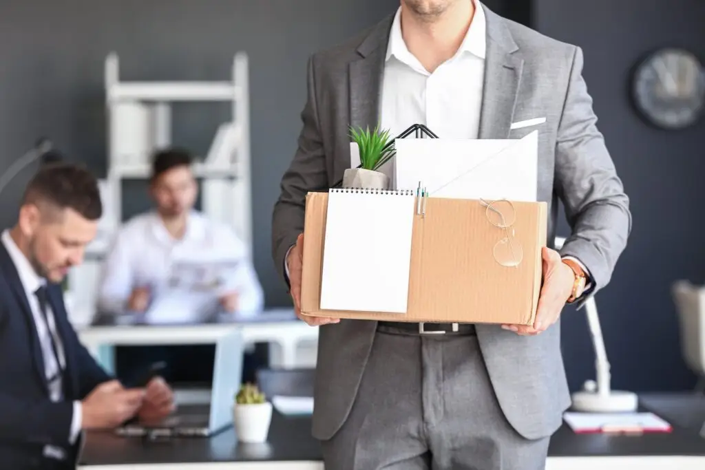 Constructively fired employee leaving the office with box of personal belongings with two current employees working in the background