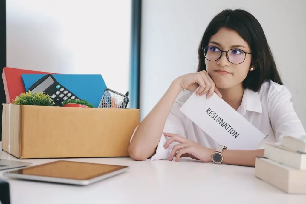 Young employee holding her resignation letter next to box of personal belongings at her former work desk
