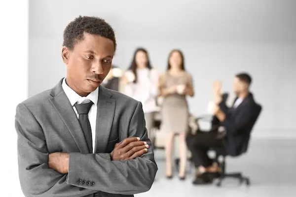 Victim of workplace discrimination in a business suit looking forlorn with three co-workers in the background in an office