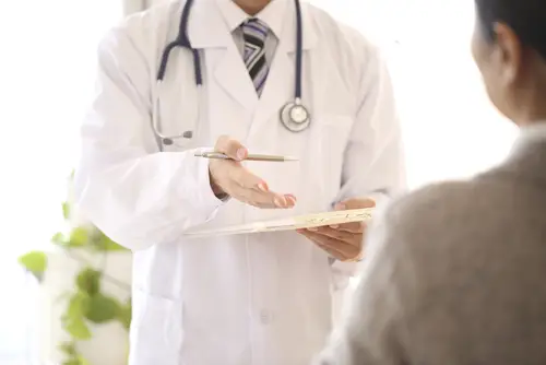 Doctor in white coat with a stethoscope and tablet talking with a seated patient with a plant in the background