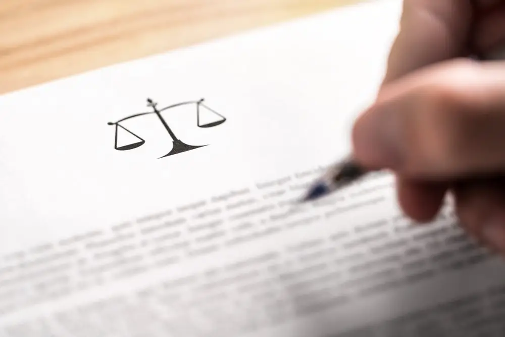 Close-up of a hand holding a pen over a legal document with a graphic of the scales of justice on the letterhead. The document is resting on a wooden table. 