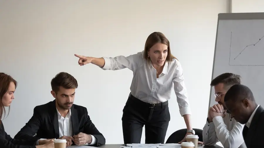 Office meeting with five employees at a board table, and the boss is standing up and instructing a worker to leave, presumably because he is African-American