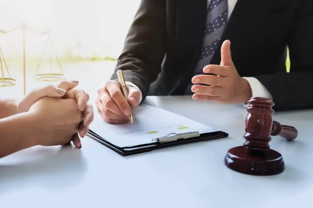 An attorney explaining employee misclassification to his client. They are sitting across from each other at a desk, and the lawyer is writing in a notebook and is near a gavel.