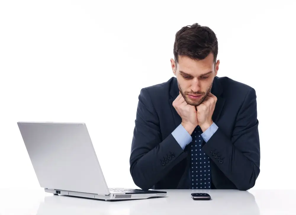 An on-call employee in a business suit at his desk in front of his laptop, and he is anxiously looking down on his phone.