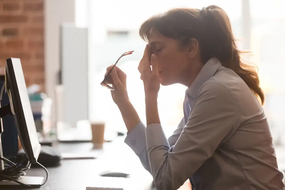 An employee feeling stress due to a workplace investigation. She is sitting at her desk in front of a desktop and rubbing her forehead while taking off her glasses. 