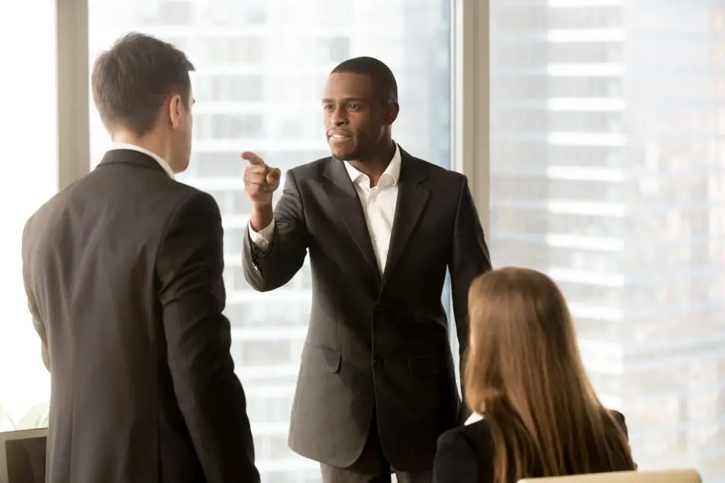 Boss pointing finger at employee during tense office meeting with another co-worker, with a floor-to-ceiling window in the background