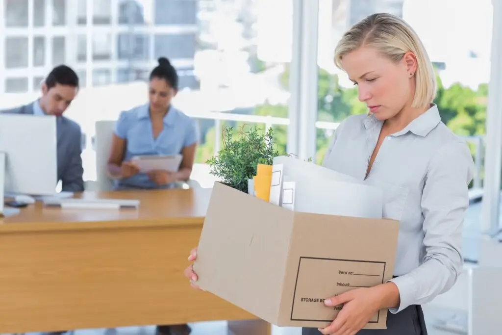 Sad-looking fired employee holding box of belongings as she leaves the office, where two other employees remain at a desk