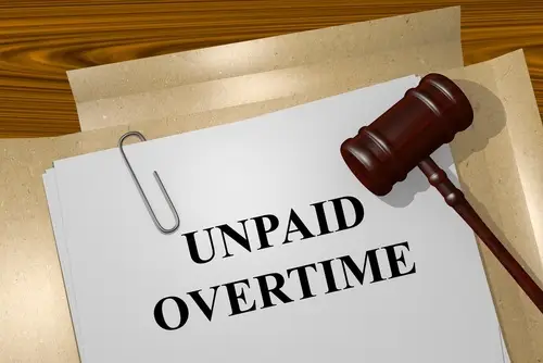 A stack of white paper paper-clipped together, and the top page reads "Unpaid Overtime." The papers are resting on a manila folder on a wooden table, and there is a gavel on resting on the paper