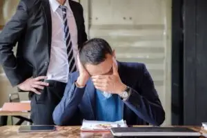 An employer at his work desk holding his head in his hands in despair while his employer stands behind him berating him, with a window in the background