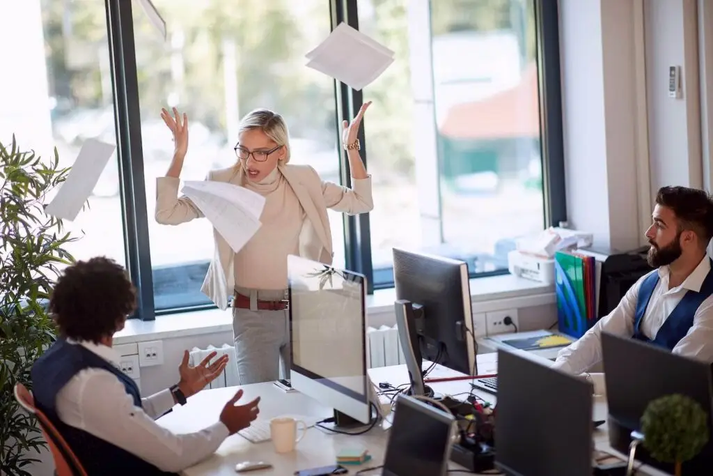 Manager in an office throwing papers in the air in anger as two other employees, who are seated at their desktop computers, look on worriedly, with an office window in the background