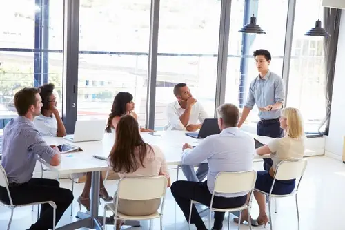 Group of eight office workers meeting around a board table, and one of them is standing at the head of the table and addressing the others. Floor-to-ceiling windows are in the background, overlooking a cityscape.