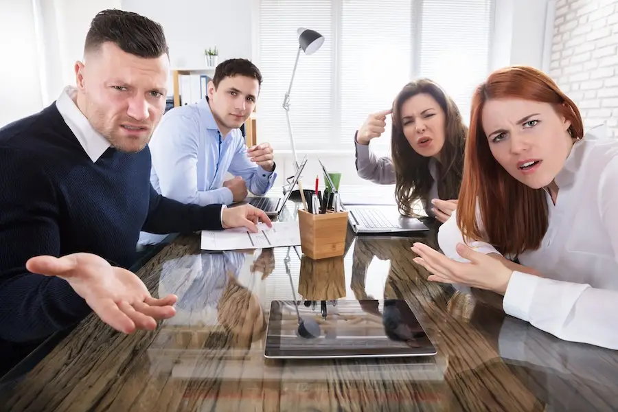 Four angry, bullying co-workers at a board room desk with tablets and a lamp looking towards the camera, with an office window in the background