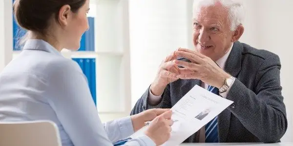 Young female employer interviewing an elderly male job candidate in a suit and tie. She is holding his resume while asking him questions over her office desk.