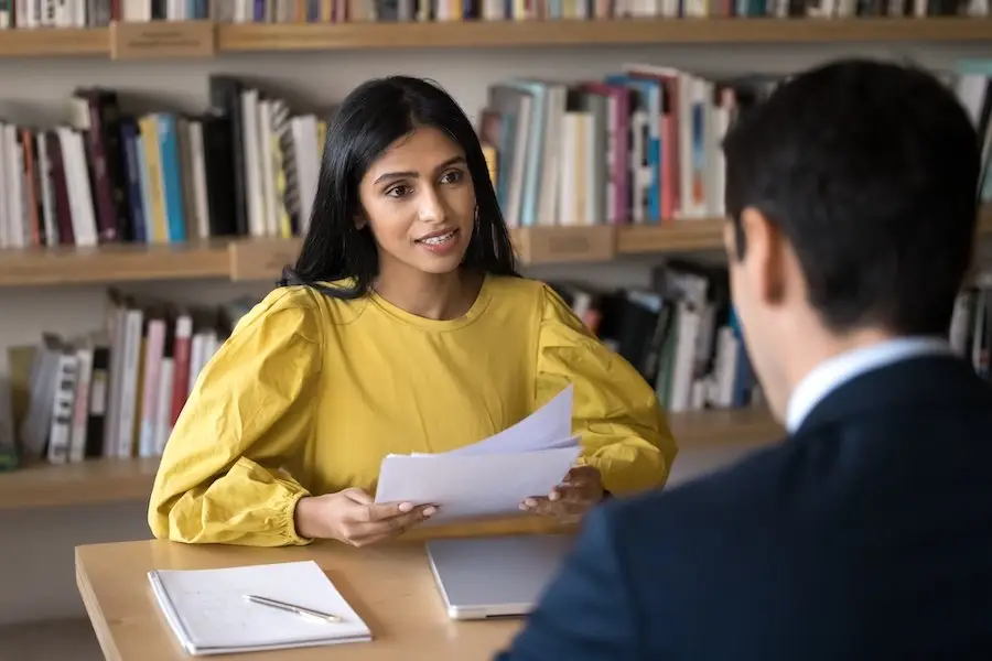 A job applicant sitting across an office desk from an employer, and she is holding copies of her resume