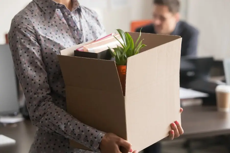 Laid off worker carrying box of belongings out of office as her boss sits at his desk in the background