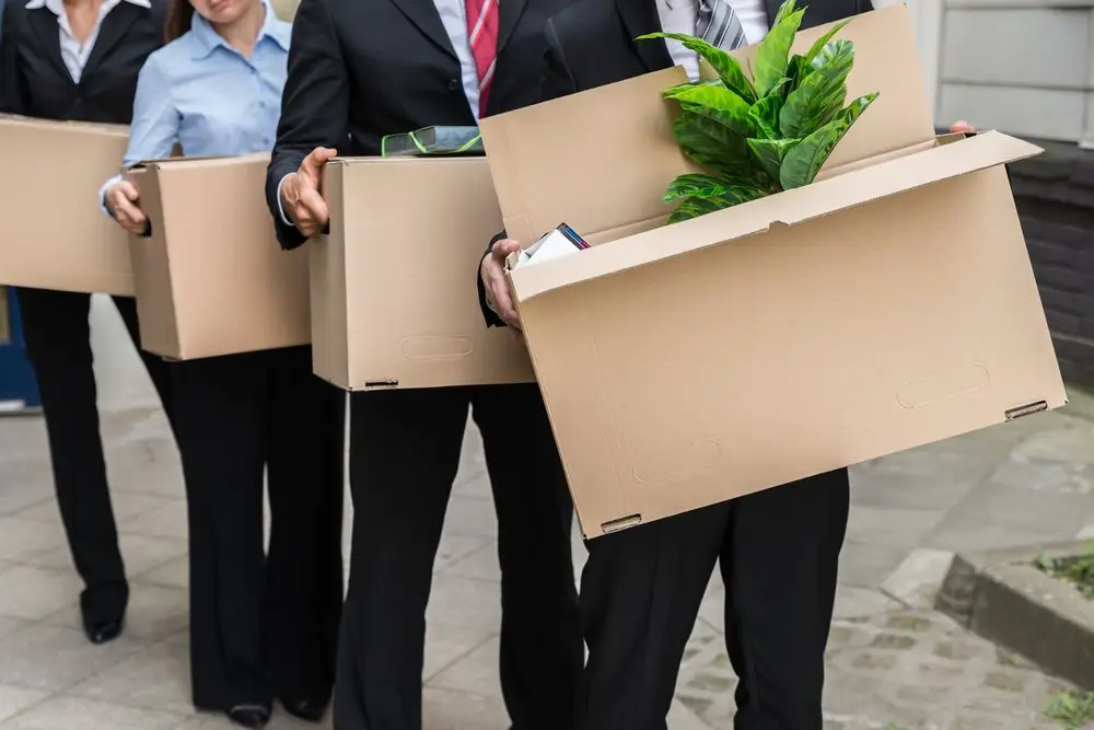 Four employees in business suits leaving an office building with their belongings in cardboard boxes after getting laid off.