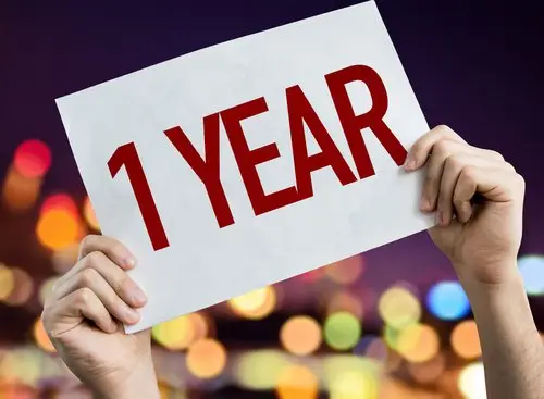 hands holding up a white sign that says "1 year" in red letters with city lights in the background