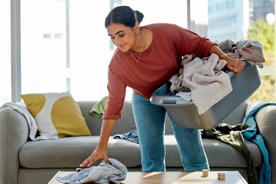 Maid holding a laundry basket with one hand and picking up clothes that are strewn on a table and couch with the other