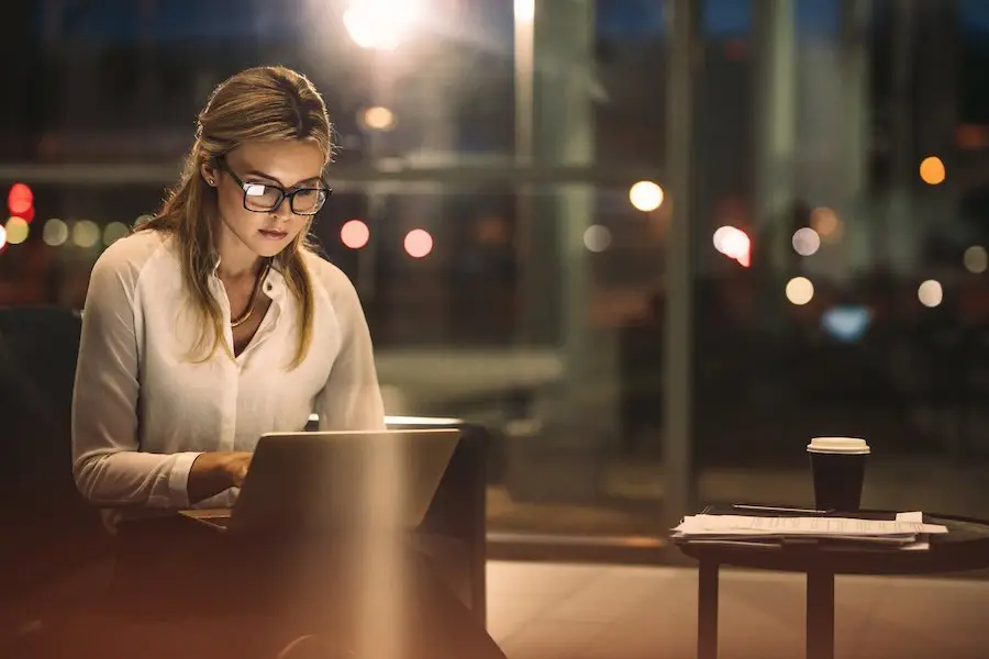 Professional typing at her laptop in her office late at night with the city lights visible through her office window
