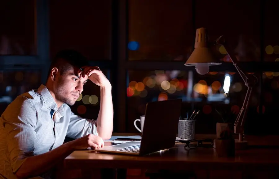 White-collar employee working late in office at his laptop, and the city lights are visible through his office window