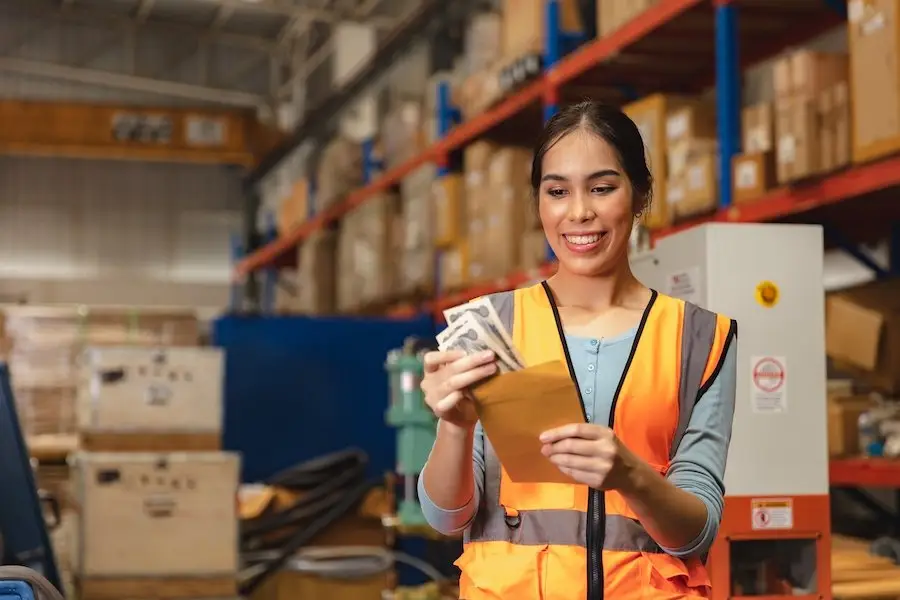 Warehouse worker in an orange vest looking happy as she takes cash out of an envelope, which is her overtime pay