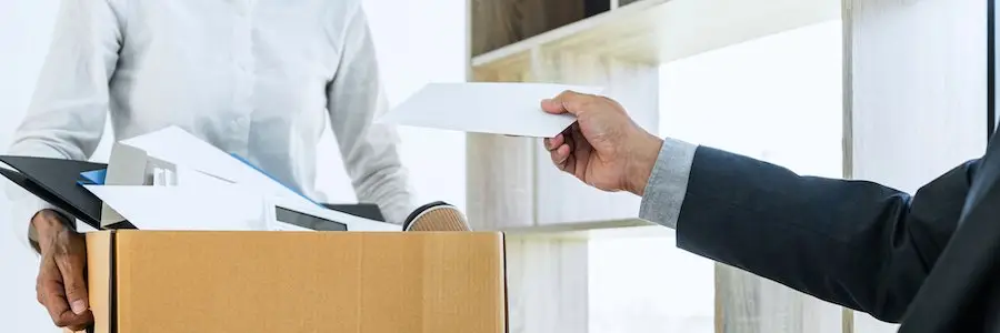 Boss in business suit handing final paycheck to fired employee as he is carrying a cardboard box of his belongings out of the office