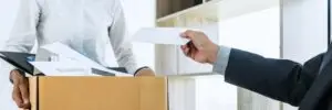 Boss handing final paycheck to fired worker holding box of belongings 