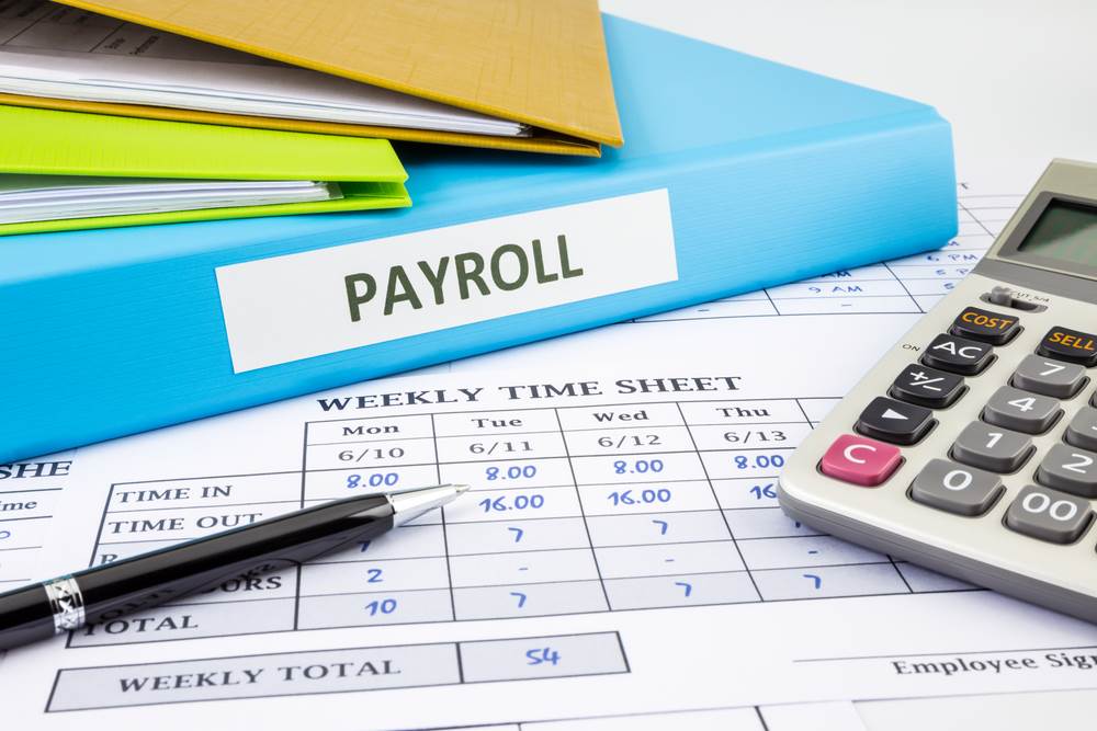 Weekly time sheet on a desk along with a pen, calculator, and a blue binder with "payroll" on the spine.