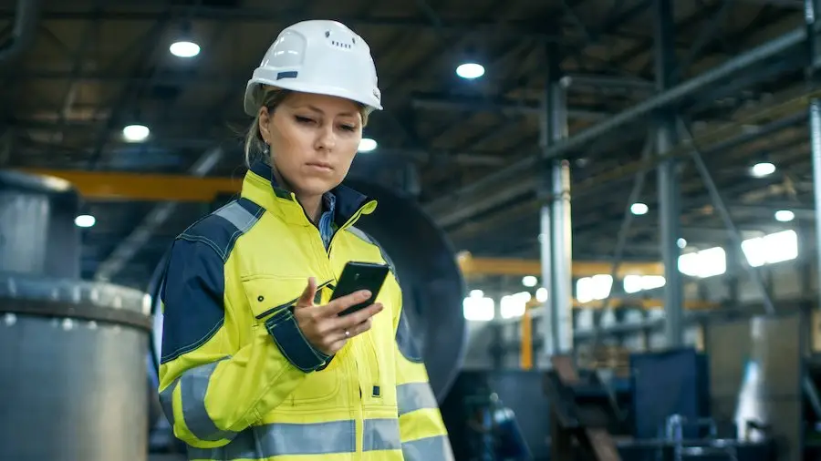 Female worker in a white hard hat and yellow jacket in a warehouse holding a cell phone 