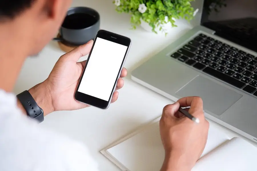 Over-the-shoulder shot of an employee holding a cell phone at his desk behind a laptop, and he is writing on a notepad with the other hand