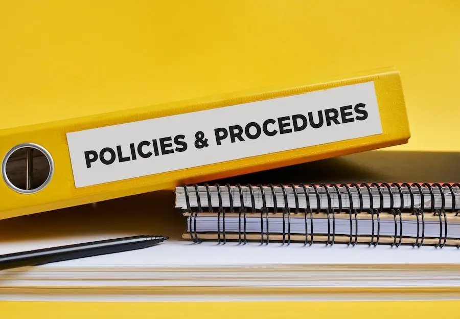 Yellow binder with the label "Policies & Procedures" along with a pile of paper and two spiral notebooks