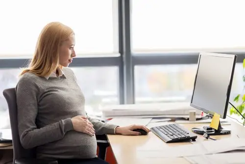 Pregnant employee holding her belly and looking at her computer on her desk in her office with a floor-to-ceiling window in the background