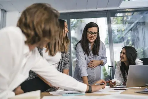 Office meeting of four women looking at papers over a desk, and one of them is holding her pregnant stomach while her co-workers smile