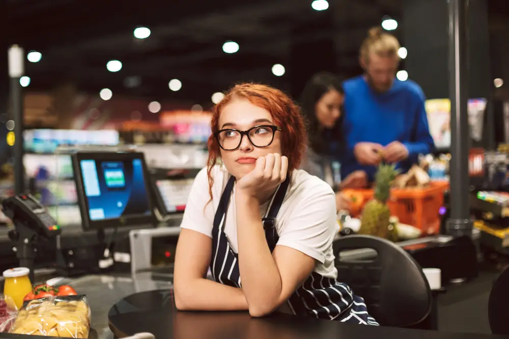 A young retail worker in an apron at her check-out station looking bored due to the lack of activity at work. 