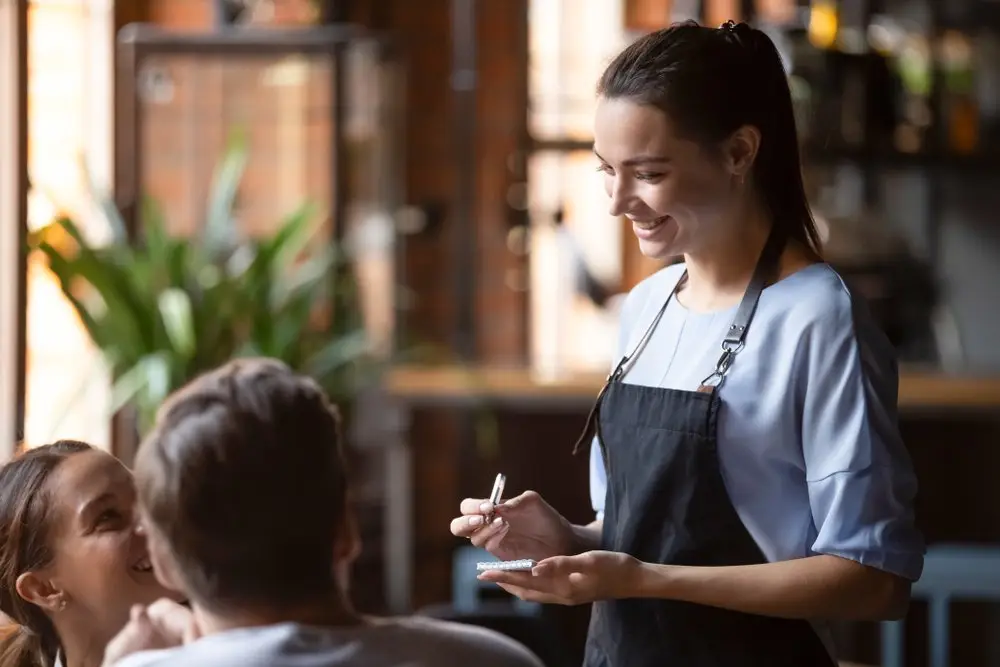 A smiling young server in an apron at a California restaurant taking down an order from a young couple at a table