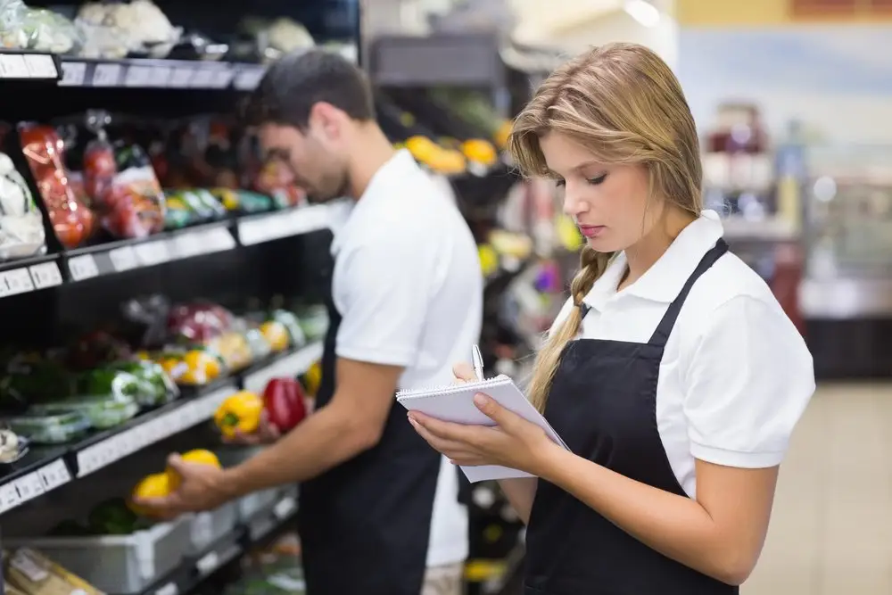 Two grocery workers with aprons stocking shelves. One is holding bell peppers while the other is taking notes.