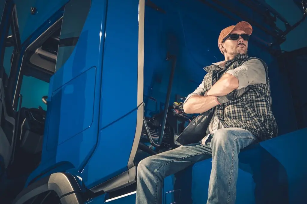 Truck driver in sunglasses and a ball cap folding his arms while taking a break outside of his truck