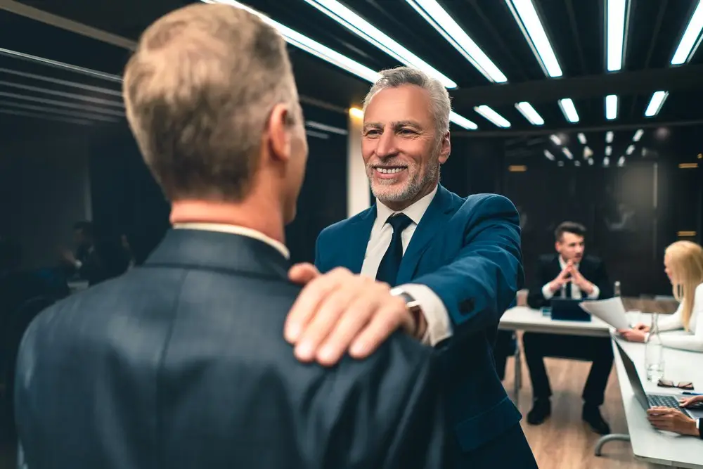 A boss in a business suit rests his hand on the shoulder of a former employee in a goodwill gesture, and two co-workers are in the background