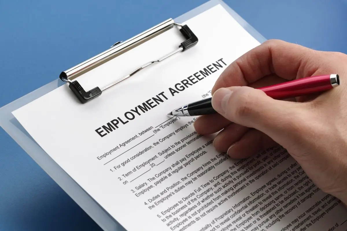 Man holding a pen filling out an employment agreement on a clipboard against a blue background