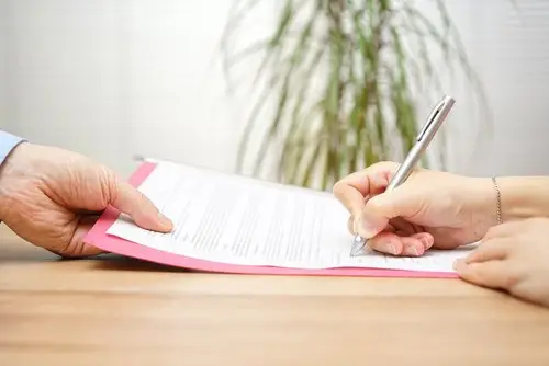 Close up of a boss handing a severance contract to a laid off employee, who is signing it with a silver pen. The contract is resting on a wooden table with a plant in the background. 
