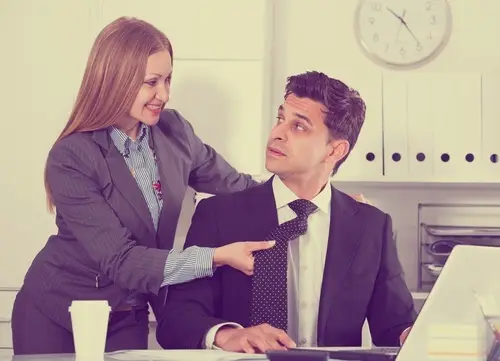 Female boss in a business suit harassing an annoyed male employee by touching his back and holding his tie in a suggestive manner