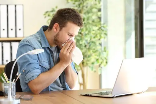 Man blowing nose in front of a laptop, who should be entitled to paid sick leave in California